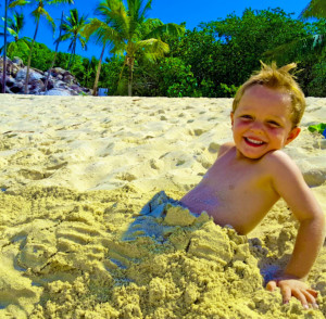 Happy young boy buried in the sand smiling, enjoying a family vacation with our private air charter to the BVI